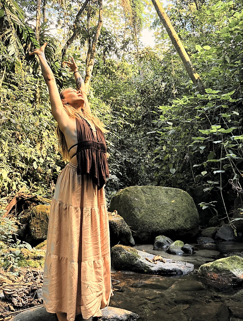 Skye standing barefoot on a rock in the stream, one arm reaching up into the canopy.