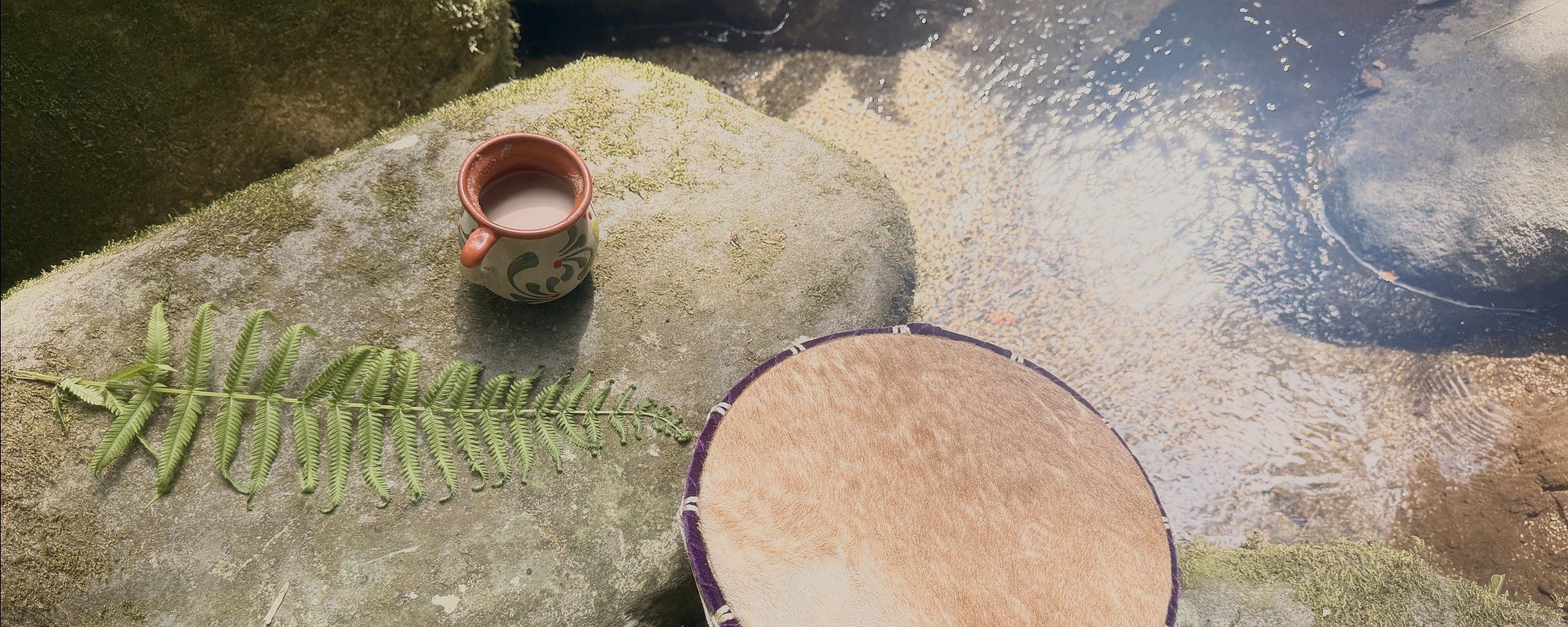 A frame drum, a clay cup of cacao and a fern frond resting on a mossy stone beside running water.