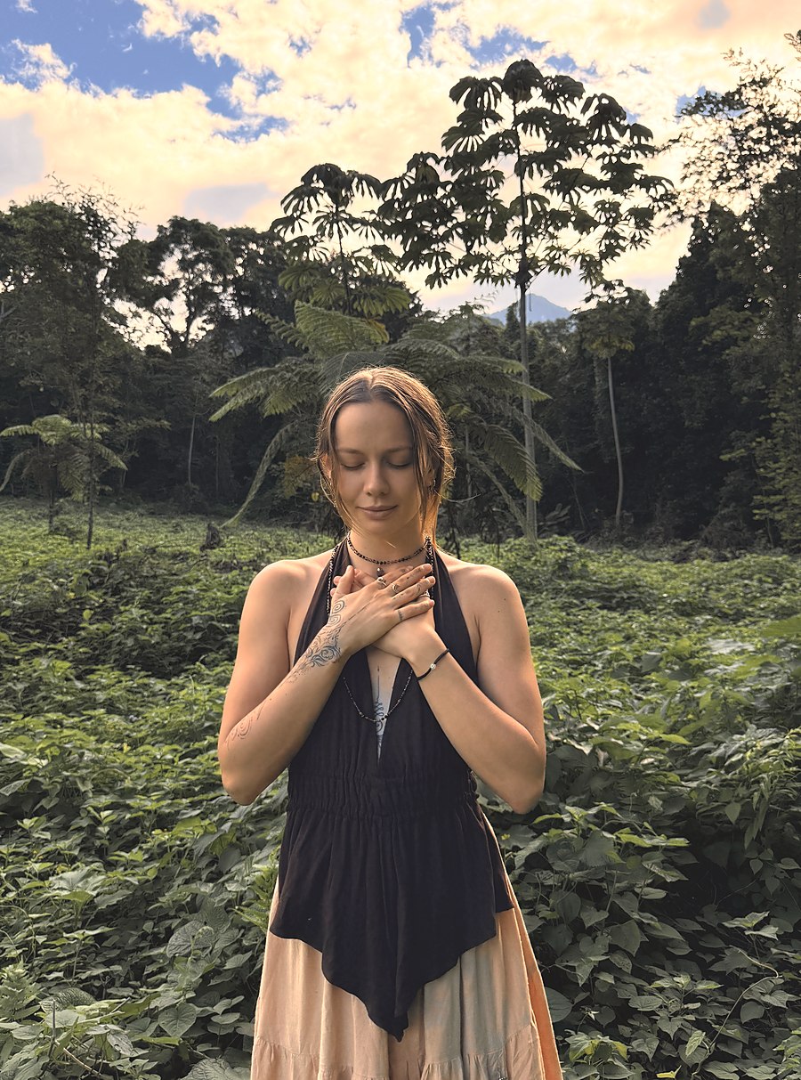 Skye standing in a jungle clearing with both hands resting over her heart, eyes closed.