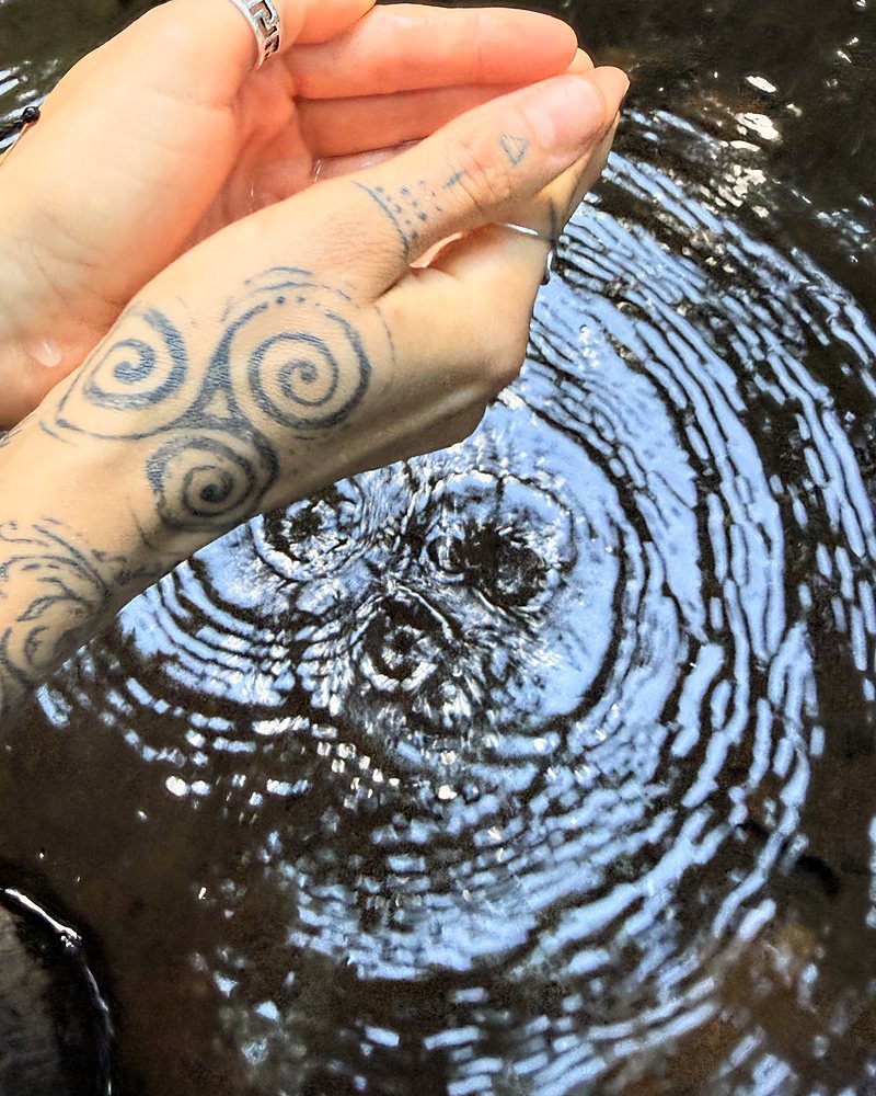Cupped hands lifted from a stream, a triple spiral tattooed on the wrist, rings spreading across the water.